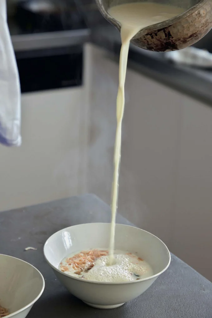 ベースの材料を入れた器から約30cmの高い位置から、温めた豆乳を注ぎ入れる。