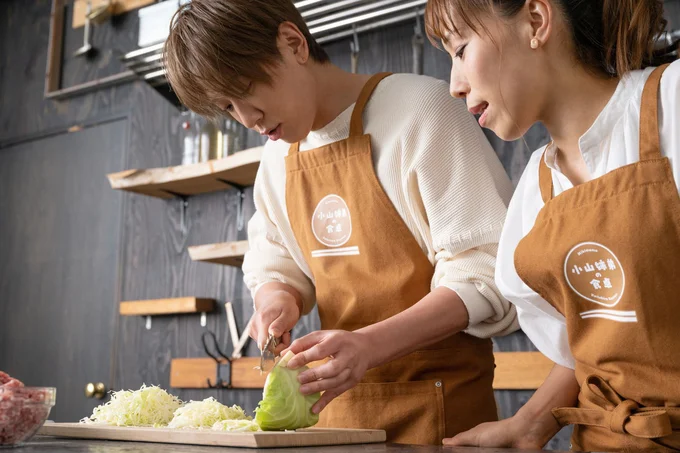 手早くできるキャベツの千切り方法を、姉からレクチャーされる小山慶一郎さん（▷二人の料理の様子は次からも続く）