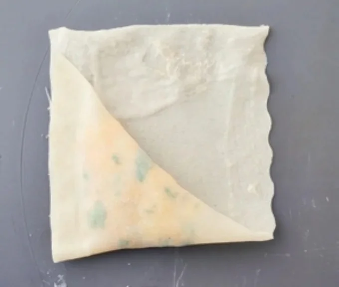 『三角春巻きの包み方』② 奥側に折り畳み、さらに三角形になるように折り、重なった辺どうしを指で押さえて密着させる。