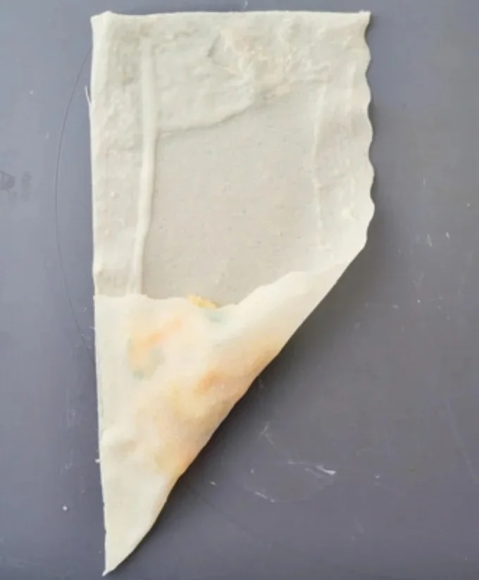 『三角春巻きの包み方』①　春巻きの皮(半分に切ったもの)を縦長に置き、皮の下半分の中央に具材をのせる。 手前以外の 3 辺にのりをぬる。具材がはみ出さないよう、手前の辺を左の辺に重ねるように折り、重なった辺どうしを指で押さえて密着させる。