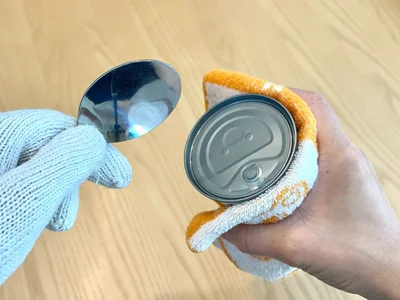軍手をはめた手でスプーンを持ち、タオルを持った手で缶詰を押さえる（▶次へ）