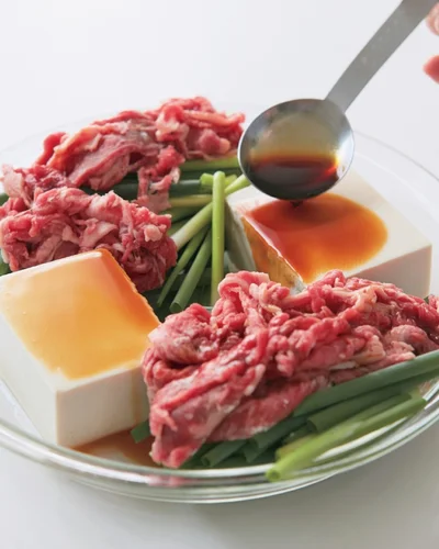 左右に豆腐を置き、中央以外のあいているところに万能ねぎを広げ、小麦粉をまぶした牛肉を均等に乗せる。「しみしみレンジ肉豆腐」