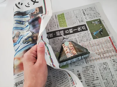 新聞紙でおにぎりを優しく包みます（▶次へ）