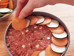 ケーキ型に半量のミートローフだねを平らにびっしり詰め、トマトを重ならないように並べる。さらに残りのたねを詰め、表面に野菜を飾る。