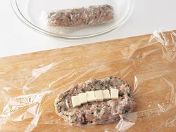 ラップの上にひき肉だねを広げ、チーズを置いて棒状に形を整える。肉汁が出るので耐熱皿に入れて加熱する。