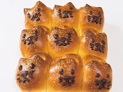【猫アレンジも！】「ごまとプレーンのちぎりパン」の生地にごまはまぶさず、全部プレーンの生地にする。牛乳をぬってから、キッチンばさみで上部２カ所に浅く切り込みを入れ、同様に焼く。さめたらチョコペンで顔を描いて。